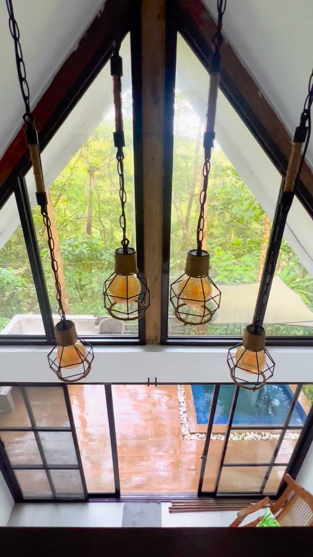 View from the La Cabaña loft interior — A-frame pendant lights hanging against the triangular glass peak, plunge pool visible below